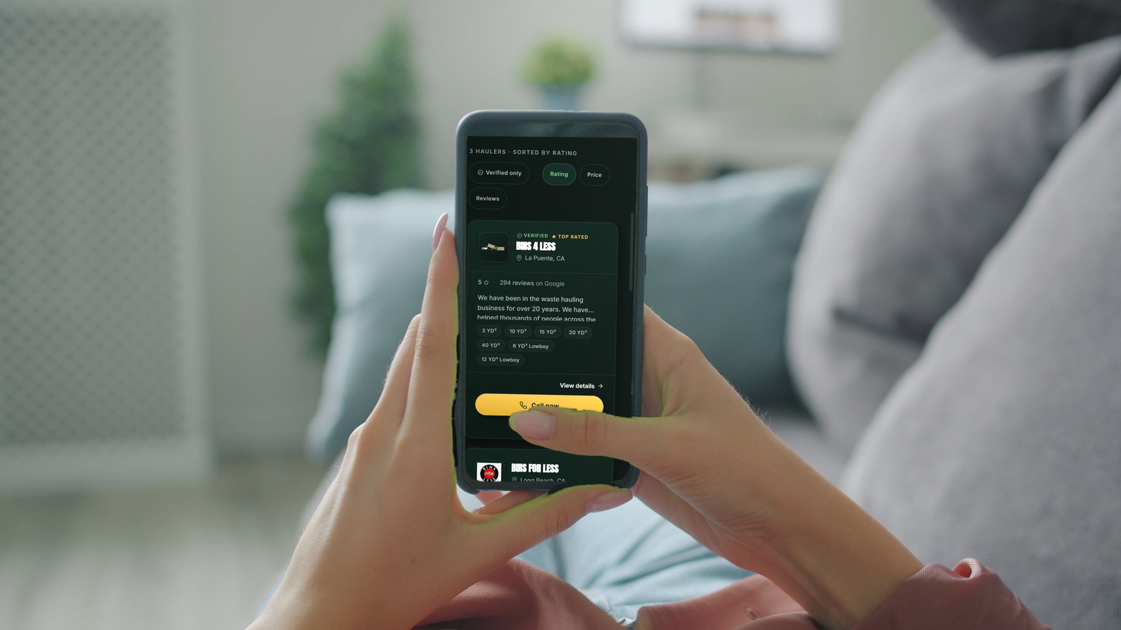 A person on a couch scrolling the Container Connect browse page on a phone, showing local hauler listings with star ratings, review counts and container sizes.