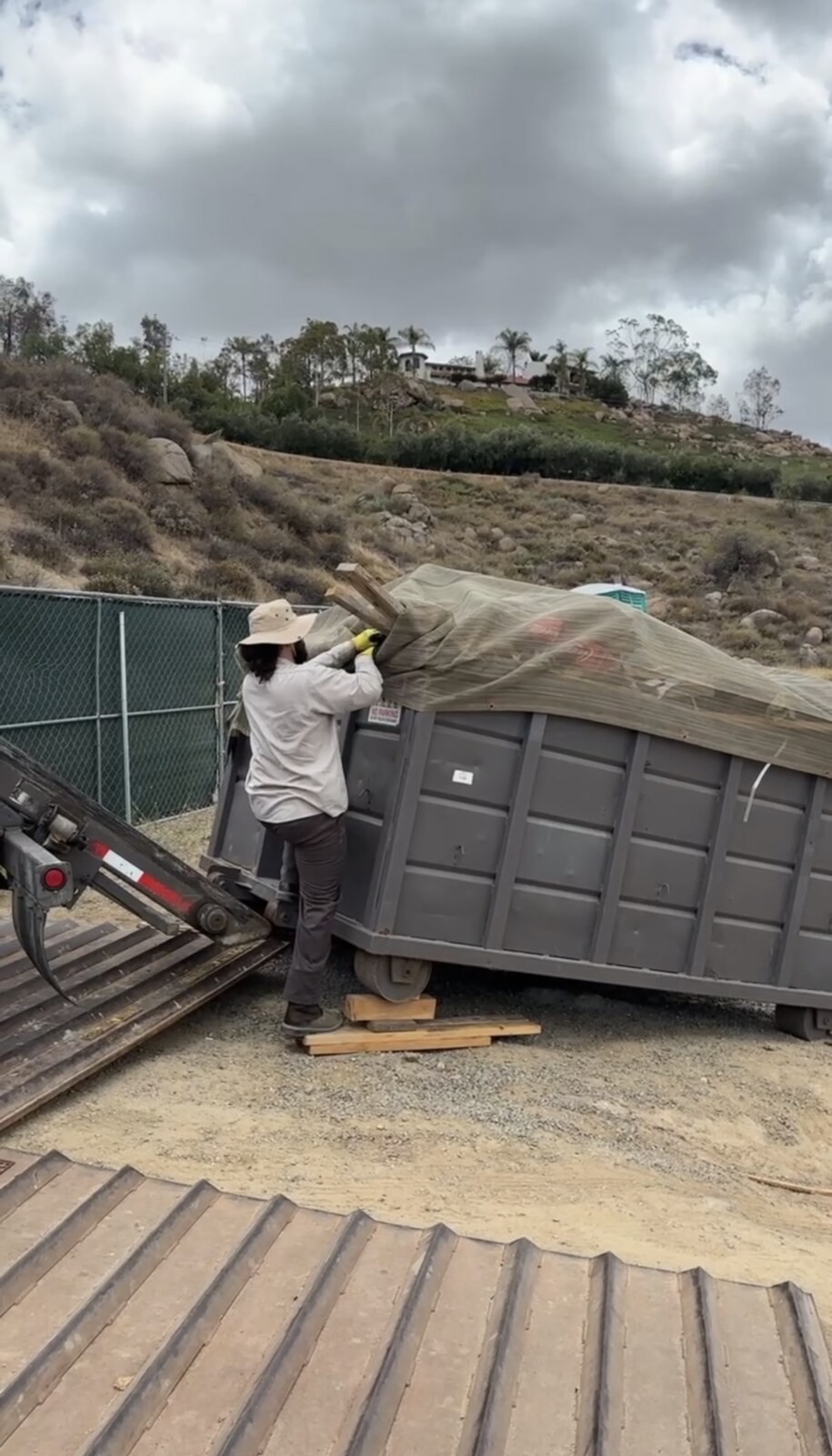 A hauling company worker in gloves pulling a tarp over a loaded roll-off container before the truck leaves the site.