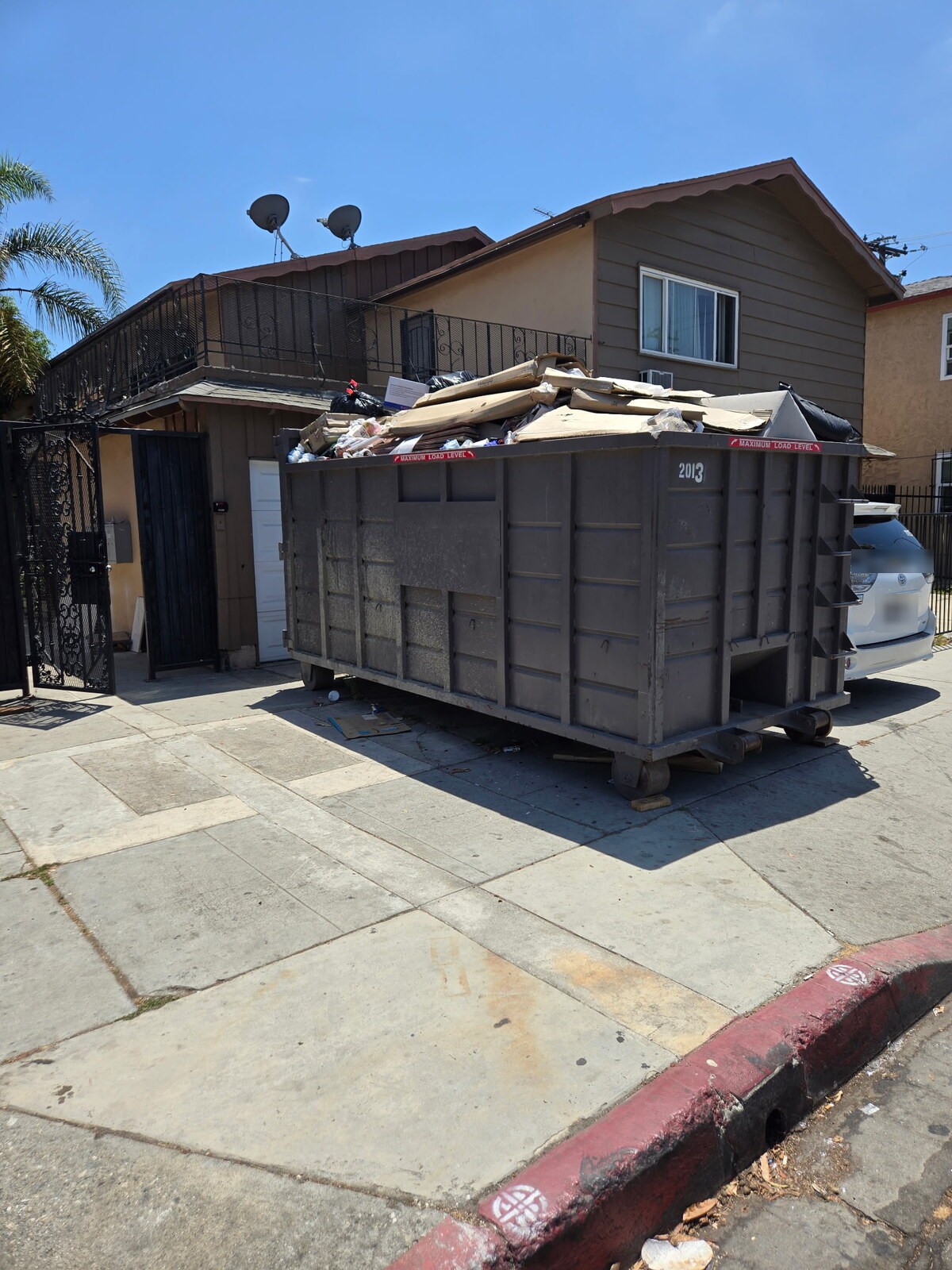 A roll-off container in a Los Angeles County driveway filled with the contents of a garage: flattened boxes, bagged waste, scrap wood and broken furniture.
