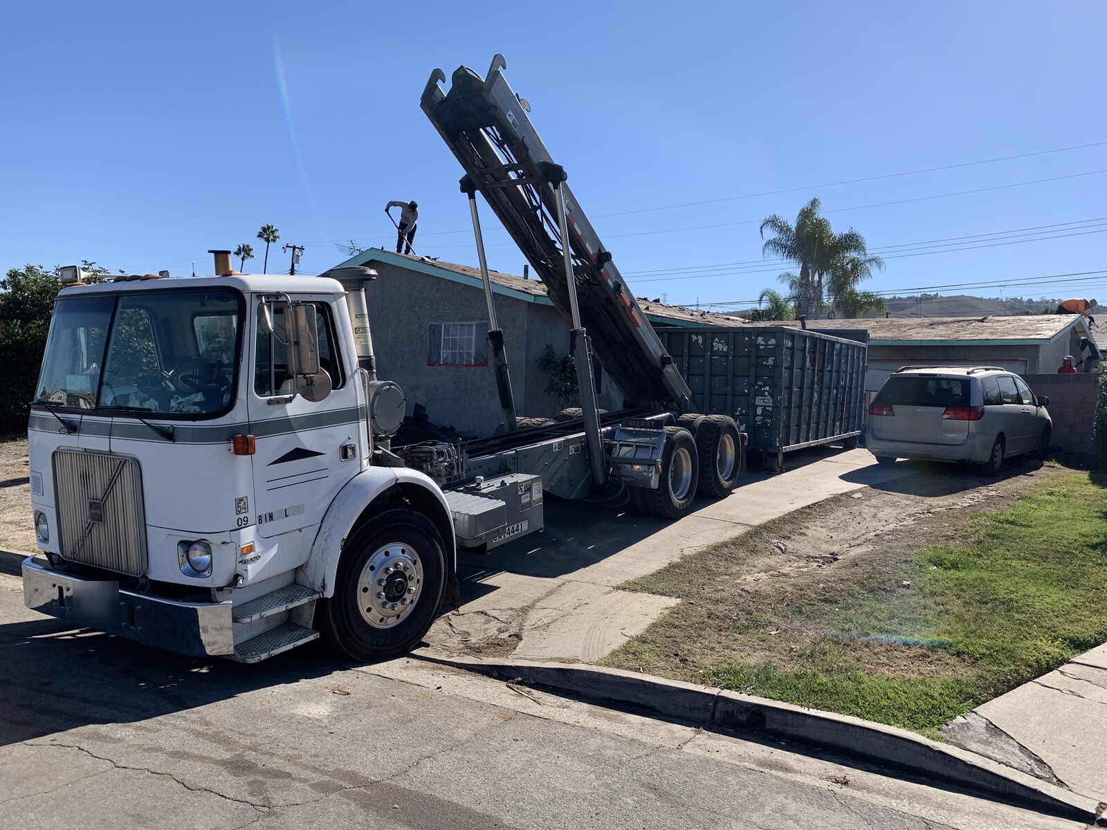 A roll-off truck with its hoist raised, setting a roll-off container down beside a Los Angeles County home during a roof tear-off.